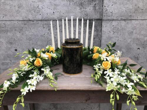 The Watering Can | A semi circular yellow and white floral arrnagement with a row of tall white tapers placed behind the urn.