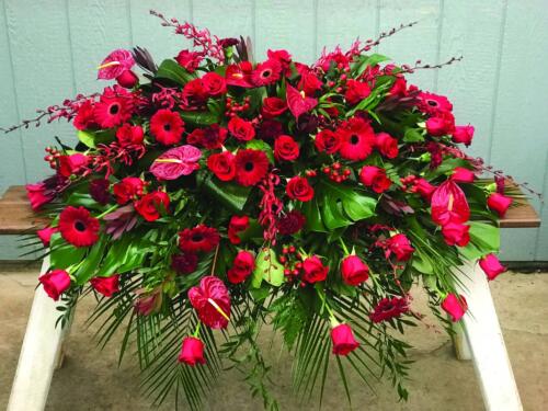 The Watering Can | An all red casket spray made with roses, leucadendron, gerbera daiseies, anthuriums, carnations, annabelle orchids, and hypericum flowers in a lush bed of greens..