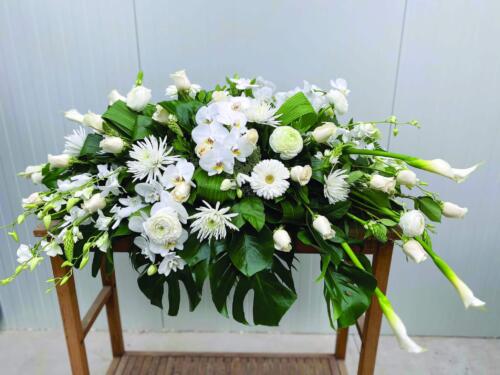 The Watering Can | An all white casket spray made with calla lilies, roses, dendrobium orchids, mums, star of Bethlehem, ranunculus, orchids, and trachelium in a bed of tropical greens.