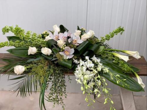 The Watering Can | An all white casket spray made with calla lilies, dendrobium orchids, white roses, bells of Ireland, cymbidium orchid blooms, hypericum in a bed of lush greens.