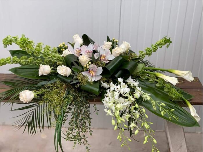 The Watering Can | An all white casket spray made with calla lilies, dendrobium orchids, white roses, bells of Ireland, cymbidium orchid blooms, hypericum in a bed of lush greens.