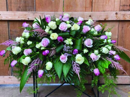 The Watering Can | A casket spray made with pink. white, and purple roses alongside astibille, waxflower and lush greens.