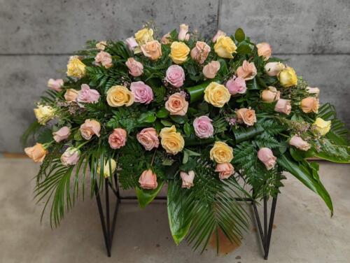 The Watering Can | A casket spray made with soft pink, soft yellow, and peach roses supported by waxflower and a variety and greens.