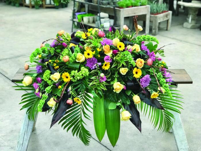 The Watering Can | A colourful caskey spray in purples and yellows made with soft yellow roses, bells of Ireland, pink lisianthus, lime green hydrangea, dark purple calla lilies, lavender mums, bright yellow gerberas, purple freesia, pink tulips, star of Bethlehem, and peach ranunculus on a bed of greens.