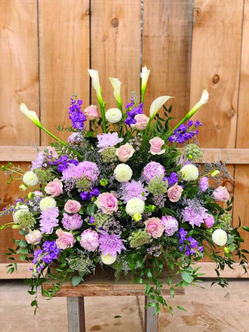 The Watering Can | A large pink and purple side spray made with white calla lilies, soft pink roses, lavender mums, purple stock, soft pink wax flower, purple limonium, white dahlias, chocolate queen anne’s lace, and soft pink peonies backed by greens.