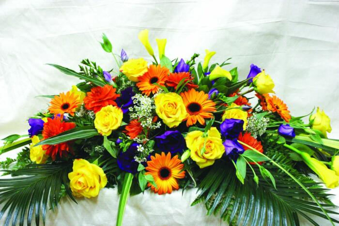 The Watering Can | A colourful casket spray made with yellow calla lilies, yellow roses, orange gerberas, orange dahlia, purple lisianthus, white wax flower, and purple purple freesia on a bed of greens.