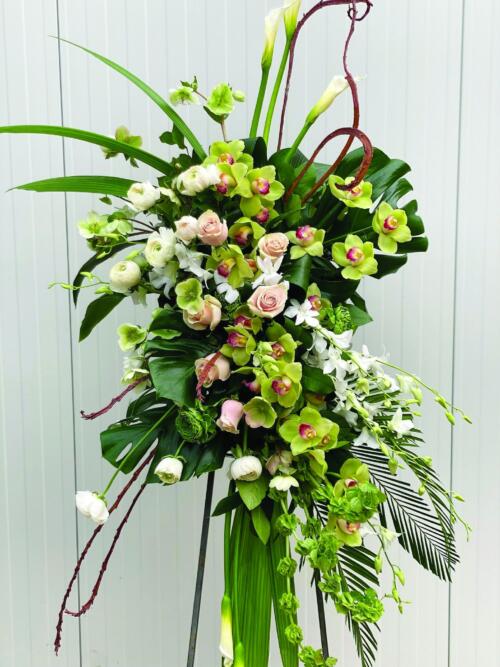 The Watering Can | A stunning modern easel spray made with white calla lilies, bells of Ireland, white & green rununculus, white helleborus, green cymbidium orchids, white dendrobium ochids, light pink roses, fantail willow, and tropical greens.