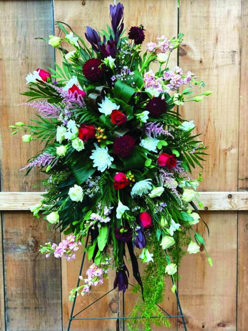 The Watering Can | An easel spray made with burgundy leucadendron, soft pink astilbe, burgundy dahlias, red roses, white mums, peach stock, peach hypericum, cream lisianthus, soft pink waxflower, white freesia on a bed of greens.