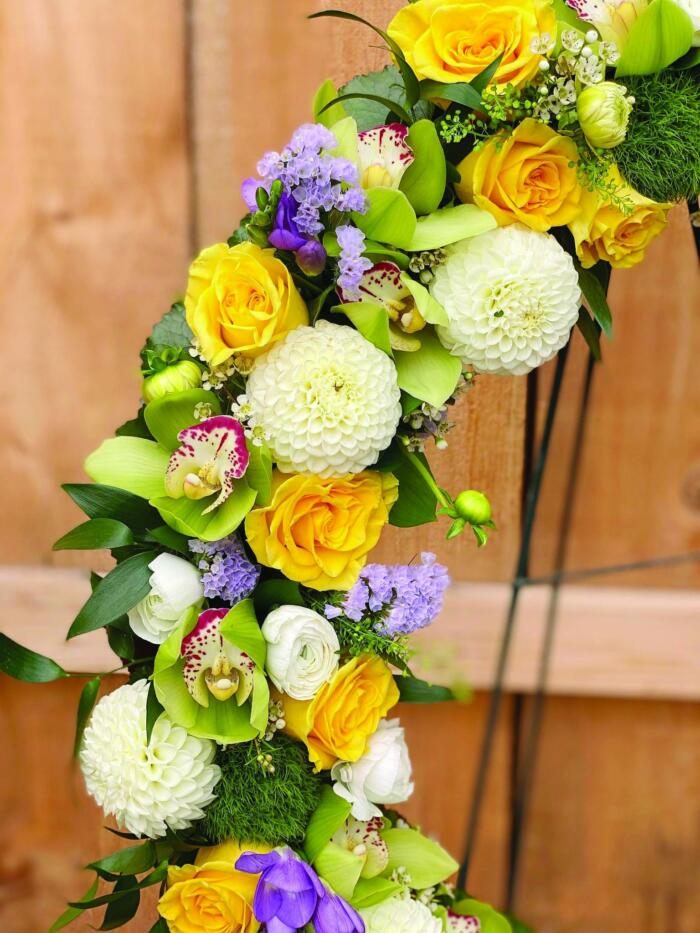 The Watering Can | Close up of the white dahlias, yellow roses, green cymbidium orchids, purple freesia, and blue statice on a floral funeral wreath.