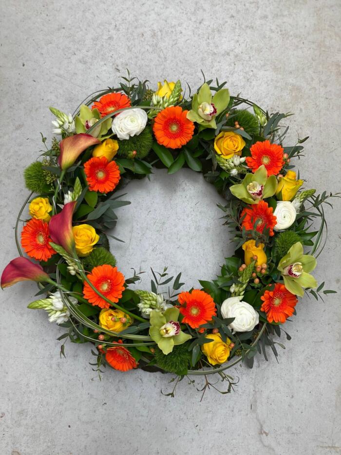 The Watering Can | A medium bevreavement wreath made with orange calla lilies, orange gerbera daisies, yellow roses, green cymbidium orchids, white ranunculus, peach hypericum, and star of Bethlehem on an abundant bed of greenery.