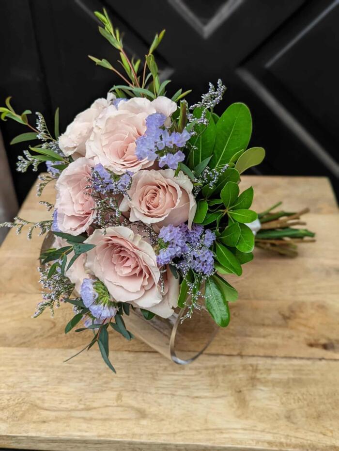 The Watering Can | A bouquet of blush roses, blue statice, limonium, and soem greenery satin wrapped lying on a table.