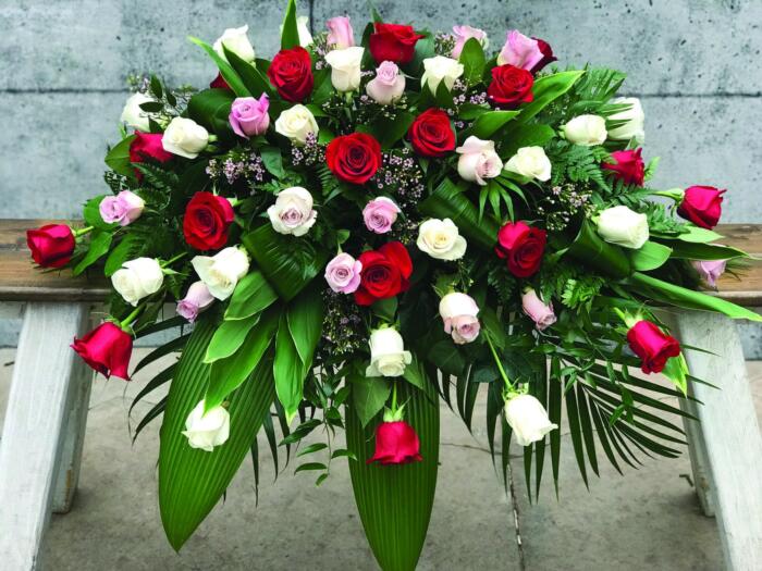 The Watering Can | A casket spray made with an assortment of red, white, and pink roses, supported by waxflower and a variety of greens.