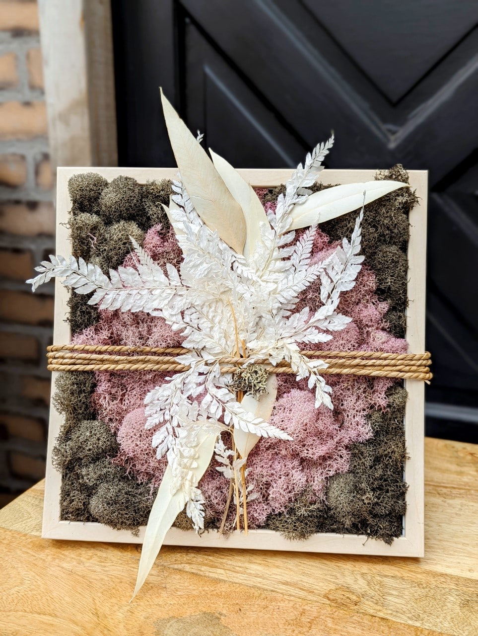 The Watering Can | A pink moss circle surrounded by brown moss in a square pale wooden frame. String is wrapped around horizontally around the moss and frame holding bleached fern and leaves.