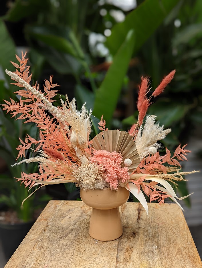The Watering Can | An European style floral arrangement using all dried goods in shades of peach and natural tones designed in a white ceramic pedestal container.
