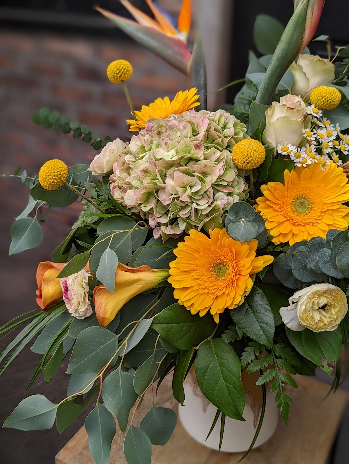 The Watering Can | Alternate view of a large asymmetrical yellow and orange bouquet in a cream, yellow, and brown ceramic vase.