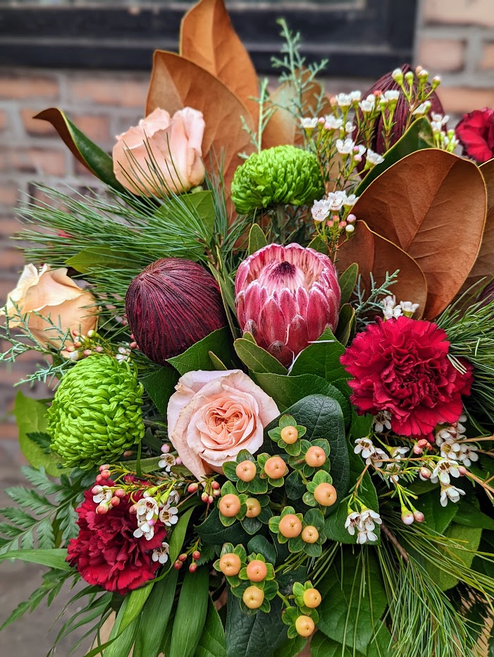 The Watering Can | Close up of protea, peach roses, peach hypericum, and green mums in a bouquet greened with magnolia, and evergreens.