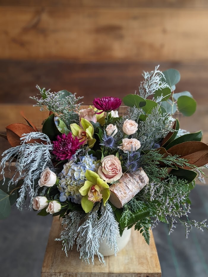 The Watering Can | A winter bouquet in vase with frosted leyland, blue hydrangea, blush roses, green cymbids, purple mums, and magnolia.